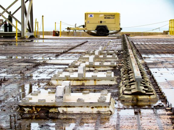 Large-format photograph: Pad Deck, Launch Complex 39-B, showing Apollo Era Crawlerway Grid Panels west of Flame Trench immediately south of the Side Flame Deflector Service/Park Positions Transport Rail. Also prominent in this image is the set of Hold-down Lugs which keep the Side Flame Deflector in place in its Service Position. Photo credit: Withheld by request.
